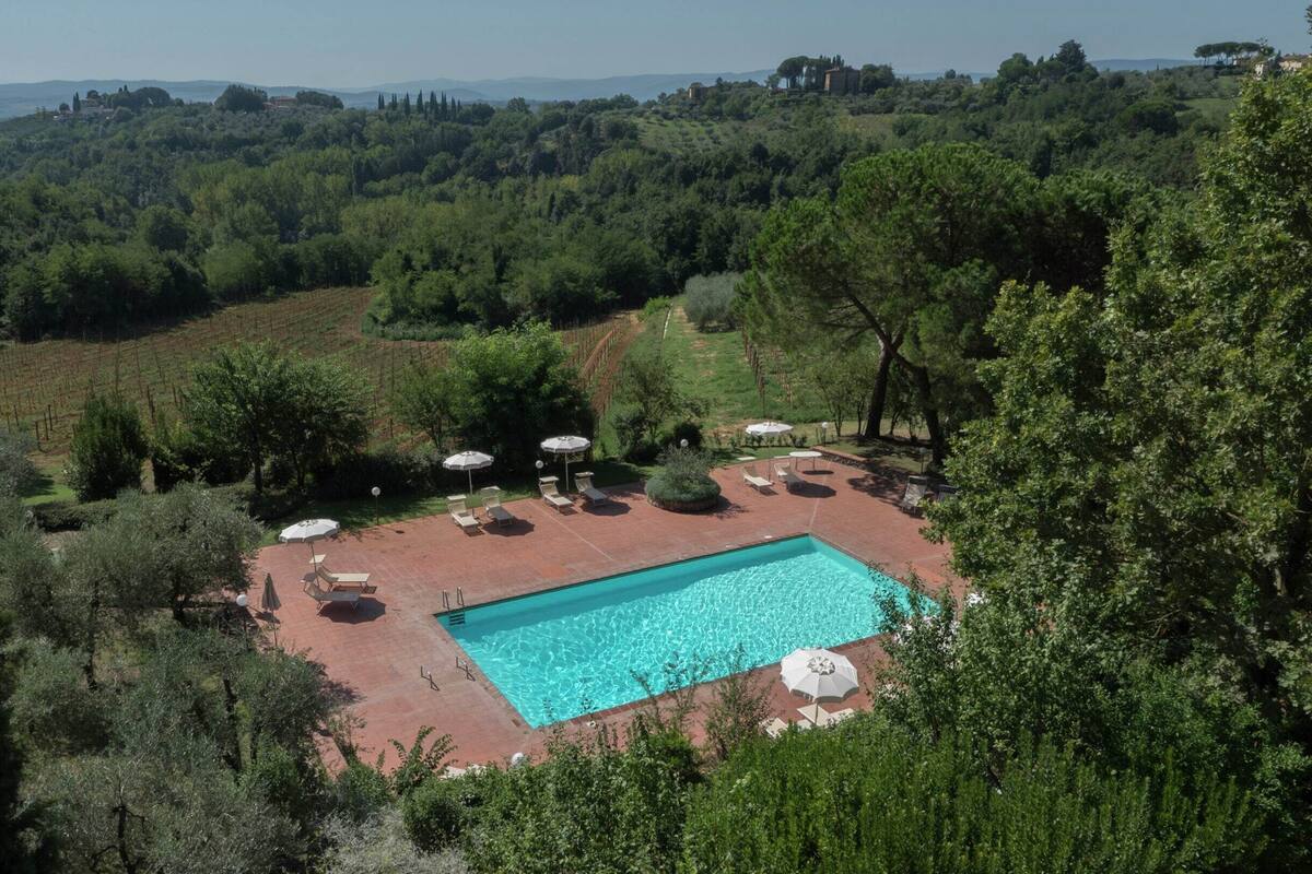 A rectangular swimming pool surrounded by a patio area with lounge chairs and umbrellas, set in a lush, hilly landscape with trees and distant buildings.