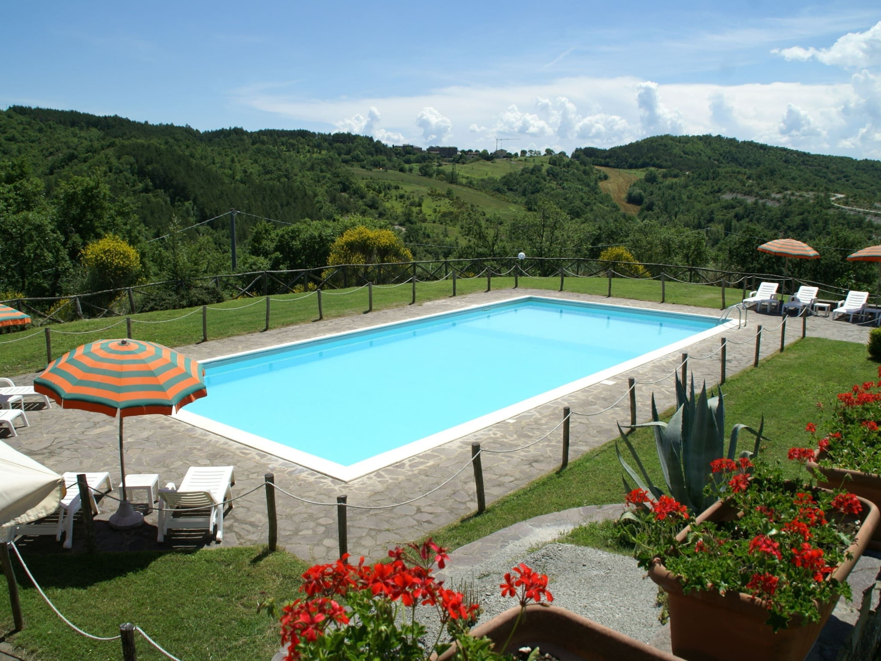 Une piscine rectangulaire bordée de pierres, entourée d’un petit muret en pierre. Des chaises longues et des parasols sont disposés le long du bord de la piscine.