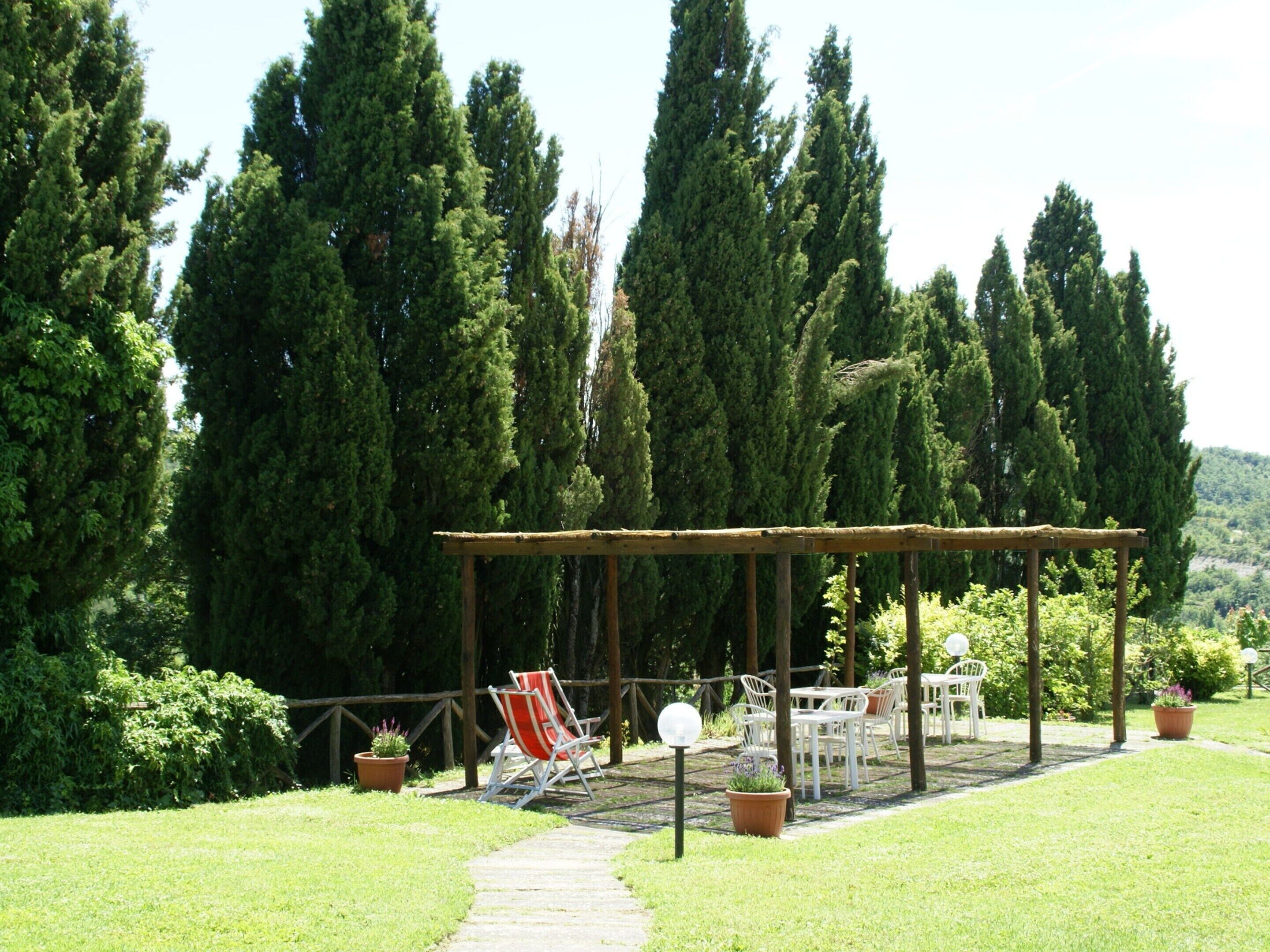 A covered outdoor seating area with white chairs and tables, surrounded by tall trees and a stone pathway.