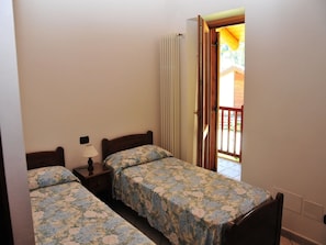 A bedroom with two single beds, a nightstand with a lamp, a radiator, and an open door leading to a balcony.