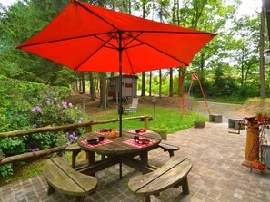 Et picnicbord med en rød parasol, omgivet af en flisebelagt terrasse og grønne planter.