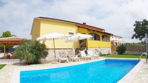 Une piscine avec des chaises longues et des parasols, un bâtiment jaune à la façade en pierre, et un ciel bleu azur.