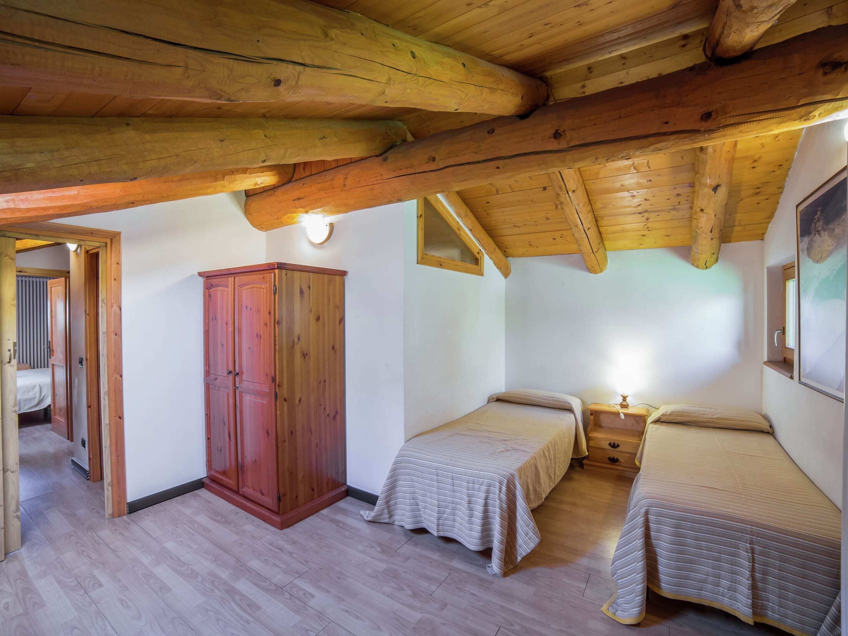 Een kamer met houten balken, twee bedden met gestreept beddengoed, een houten garderobe en een klein nachtkastje met een lamp.
