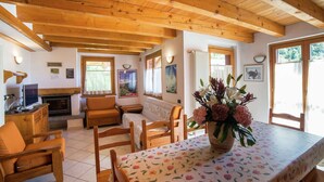 Une pièce chaleureuse avec des poutres apparentes, une cheminée, une table à manger recouverte d’une nappe à motifs floraux et une grande fenêtre agrémentée de rideaux.
