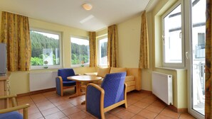 A room with a dining table, two blue chairs, a sofa, and large windows with curtains. - Ferienhaus in Willingen mit Skilift in der Nahe (Willingen)