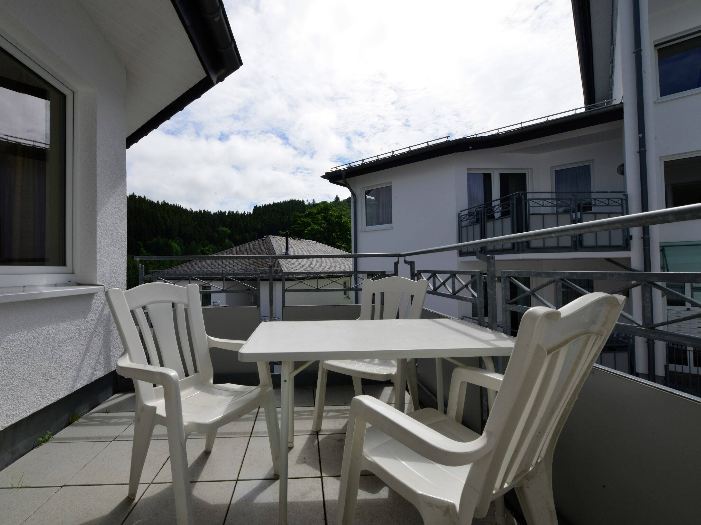 A balcony with a table and chairs, a view of a building with a balcony, and a forested hill in the background.
