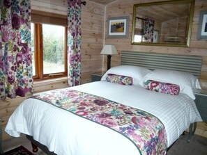 A bedroom with a bed, floral curtains, a nightstand, a lamp, a mirror, and a window with a view of trees.