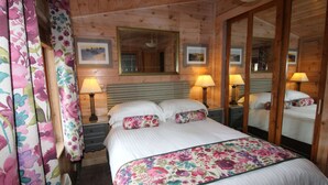 A wooden cabin room with a bed, two bedside lamps, a mirror, and floral curtains. - Lodge in Appledore Near Camber Sands Beach (Romney Marsh)