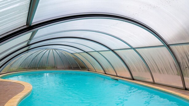 En swimmingpool med ett böjt glastak, omgiven av en terrass med kakelplattor.