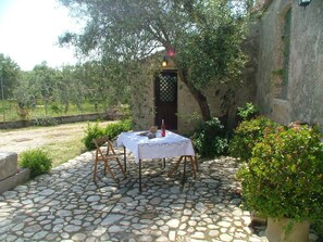 Un patio con suelo de piedra, una mesa preparada para dos personas, rodeado de vegetación y un edificio rústico.