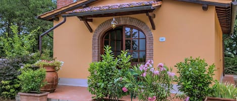 A small, single-story building with a tiled roof and a brick chimney. It features a large arched window and a door, surrounded by potted plants and a gravel driveway.
