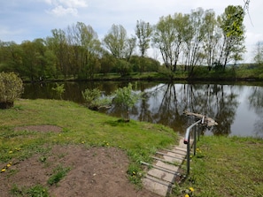A riverside path with a wooden walkway leading to a dock, surrounded by lush greenery and trees.