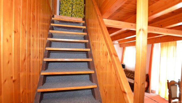 A wooden staircase with carpeted steps leading to a loft bedroom with a bed and a chair.
