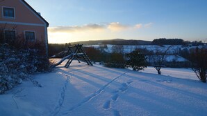 Schneebedeckte Landschaft mit einer hölzernen Schaukel und einem Haus im Hintergrund.