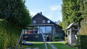 Une maison moderne dotée d’une grande baie vitrée, d’une cabane en bois pour les enfants et d’une balançoire dans le jardin.
