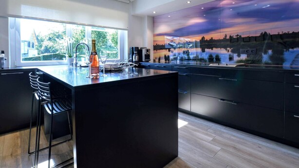 Une cuisine moderne dotée d’un grand îlot central, offrant une vue sur le coucher du soleil à travers une paroi vitrée, et une bouteille de champagne posée sur le comptoir.