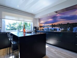 A modern kitchen with a large island, a view of a sunset through a glass wall, and a bottle of champagne on the counter. - Elsenborn Retreat Near Monschau (Buetgenbach)
