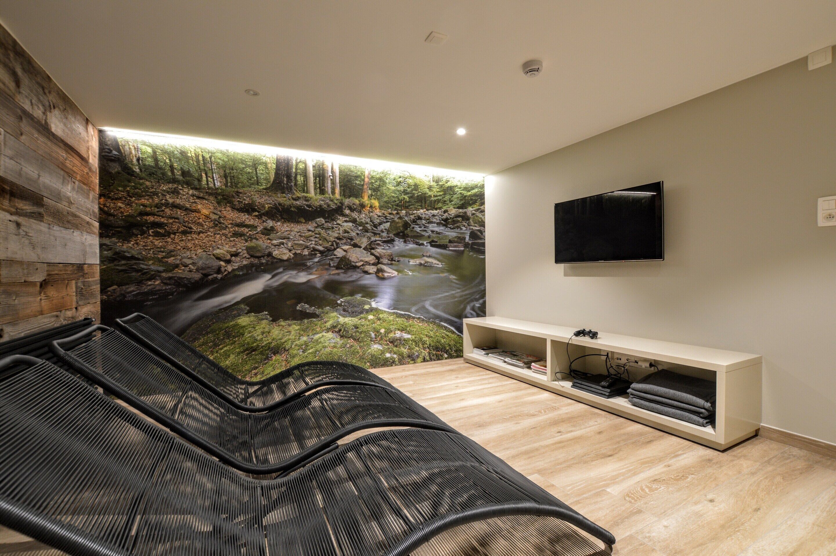 A modern living room with a large wall mural of a forest stream, a black lounge chair, a flat-screen TV, and a built-in entertainment unit.