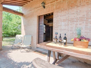 An outdoor patio with a brick wall, a wooden table with wine bottles and a fruit basket, and a white chair. - Holiday House near Lake Trasimeno (Castiglione del Lago)
