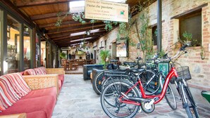 Un área de estar al aire libre acogedora con un sofá rojo y techo de madera. Se ven bicicletas apoyadas en la pared y un letrero que anuncia 'Osteria del Trasimeno Antico Borgo Pasta'.