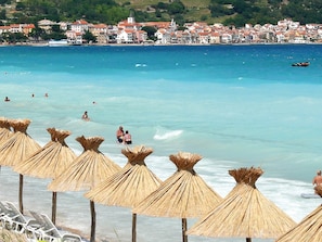 A beach with straw umbrellas and lounge chairs, a clear blue sea, and a town in the background. - Spacious Apartment in Vantacici near Sea (Malinska-Dubasnica)