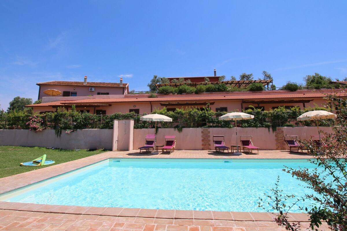 Une piscine dotée de chaises longues et de parasols, entourée d’un jardin aménagé et d’un grand bâtiment à plusieurs étages et doté de balcons.
