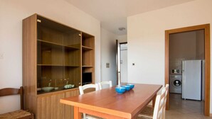 A dining area with a wooden table and chairs, a built-in wooden cabinet with shelves, and a kitchen area with a washing machine and a refrigerator. - Loving Apartment in Rosolina Mare near Venice (Rosolina)