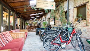 En mysig uteservering med en röd soffa och ett tak av trä. Det står cyklar lutade mot väggen och en skylt annonserar för restaurangen ”Osteria del Trasimeno Antico Borgo Pasta”.