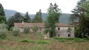 A rustic stone building with a tiled roof, surrounded by greenery and hills in the background.