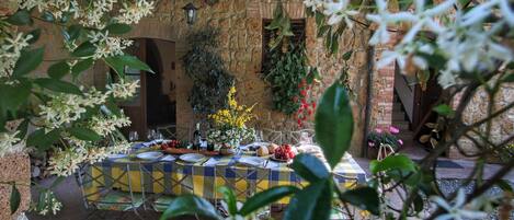 An outdoor dining area with a stone wall backdrop, a table set for a meal, and various plants and flowers.