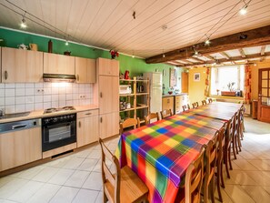 Une cuisine avec des meubles en bois, une table à manger recouverte d’une nappe colorée et une vue sur l’extérieur par une fenêtre.