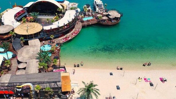 Vista aérea de um resort à beira-mar, com uma piscina grande, área de praia e palco para eventos.