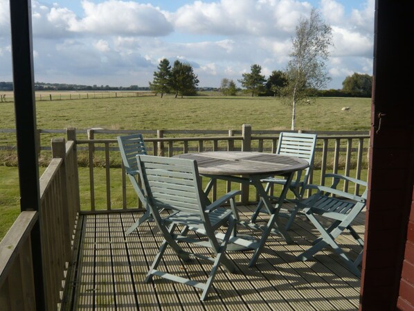 A wooden deck with a table and chairs, overlooking a grassy field. - Cosy Pine Lodge With far Reaching Country Views and Large sun Deck (Romney Marsh)