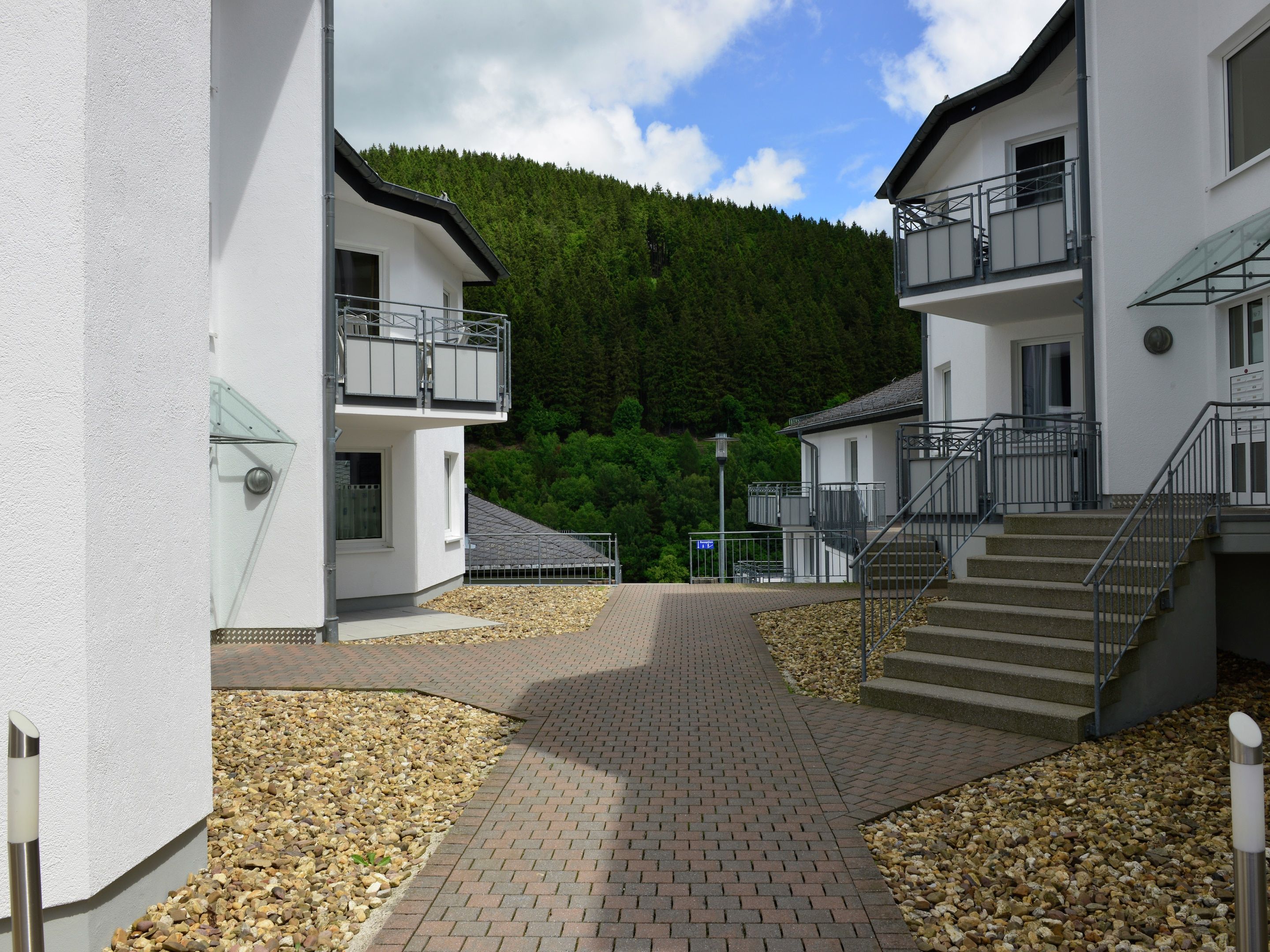 A modern building with a paved walkway, a staircase, and a view of a forested hill.