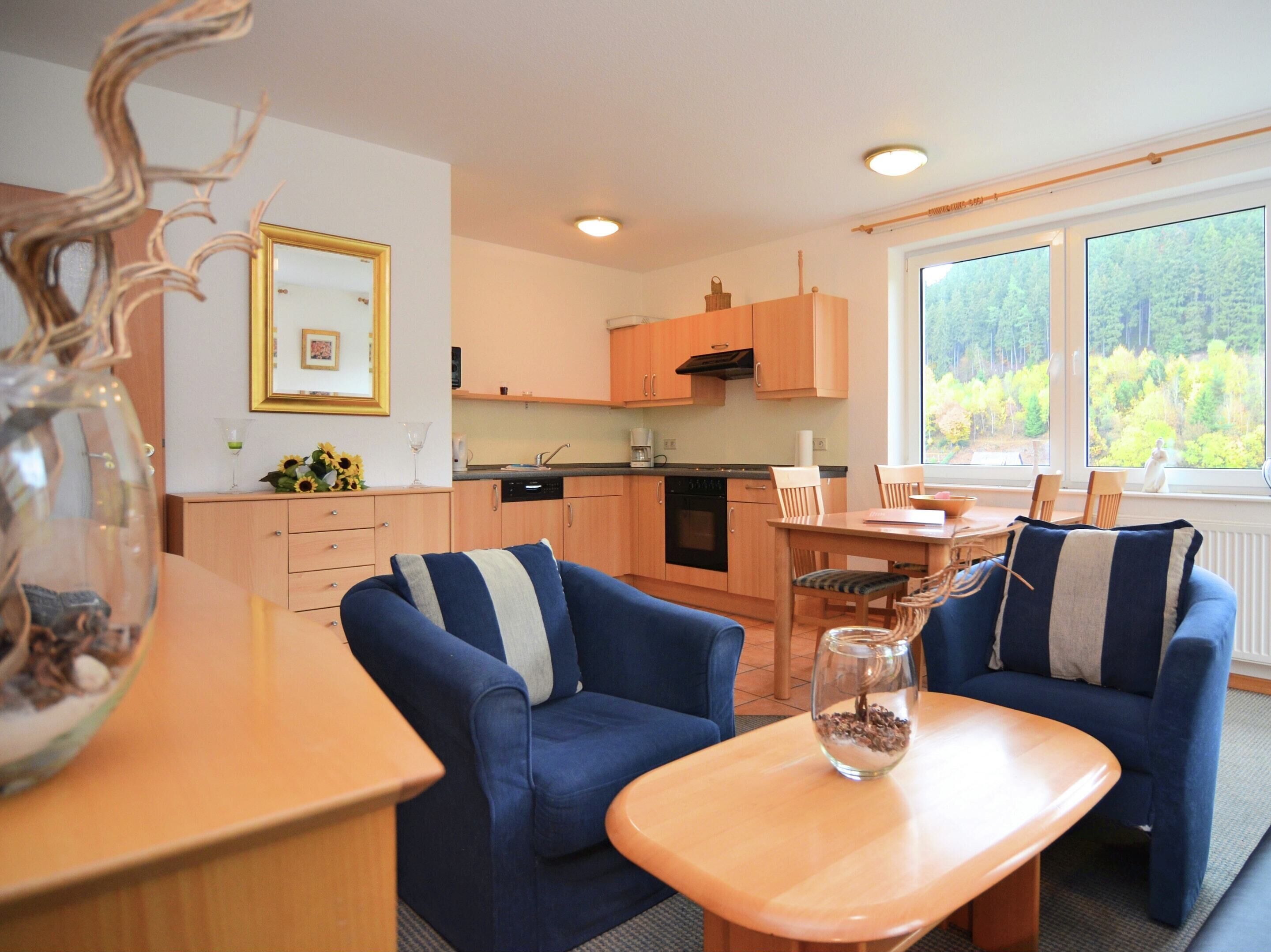 A modern kitchen with wooden cabinets, a dining area with blue sofas, and a large window with a forest view.