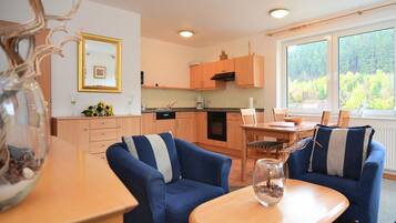A modern kitchen with wooden cabinets, a dining area with blue sofas, and a large window with a forest view.