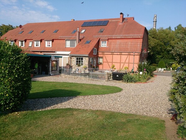 A large building with multiple windows and a sloped roof, surrounded by a gravel driveway and greenery. - Wohnung in Grundshagen mit Gartensitzplatz (Klütz)