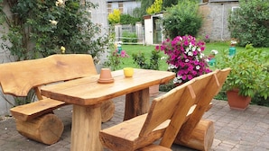 Une table et un banc en bois, installés à l’extérieur dans un jardin agrémenté de plantes en pot, avec un mur de pierres en arrière-plan.