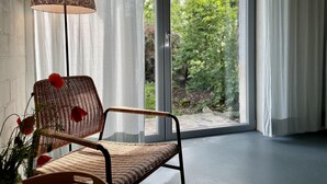Una poltrona in vimini con dettagli rossi, una lampada da terra con motivo decorativo e vista sul verde attraverso una porta in vetro.