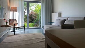 Una camera d'hotel moderna con un'ampia porta scorrevole in vetro verso un giardino, un divano, una poltrona, un tavolino e una lampada.