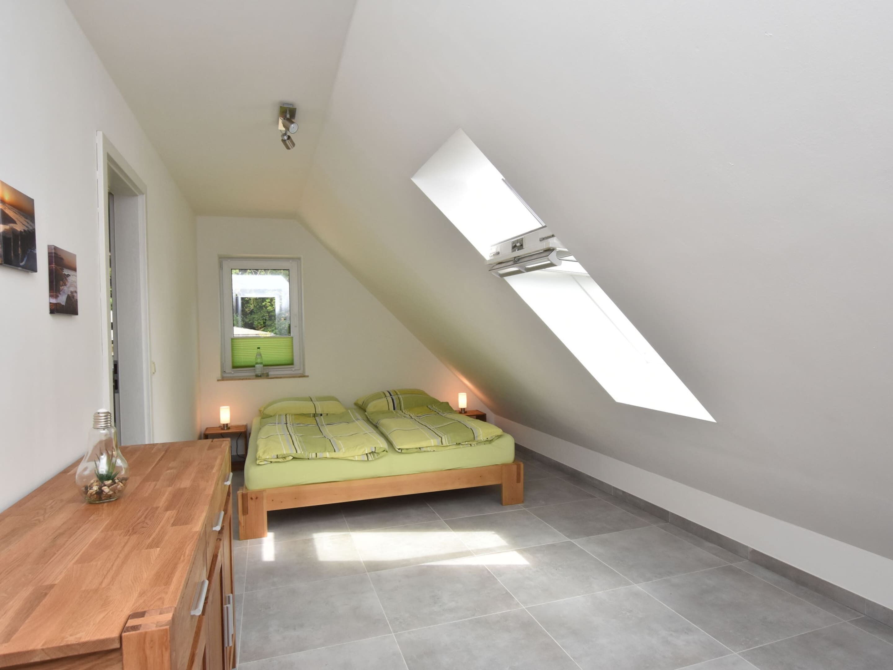 Une petite chambre à coucher moderne, avec un plafond en pente, un puits de lumière, un lit avec une literie verte, une table de chevet en bois et une petite table sur laquelle est posé un vase en verre.