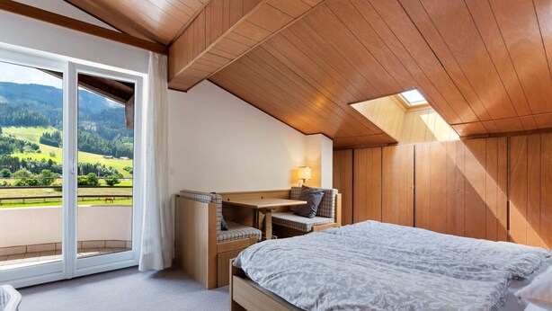 Une chambre avec un lit, un bureau et une chaise, une grande fenêtre donnant sur un paysage verdoyant, et des panneaux de plafond en bois.
