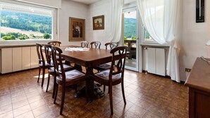 Une salle à manger avec une table et des chaises en bois, de grandes fenêtres avec des rideaux et une vue sur l’extérieur.