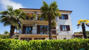 A two-story building with a balcony, surrounded by lush greenery and palm trees. - Cozy Apartment in Malinska near Sea (Malinska-Dubasnica)