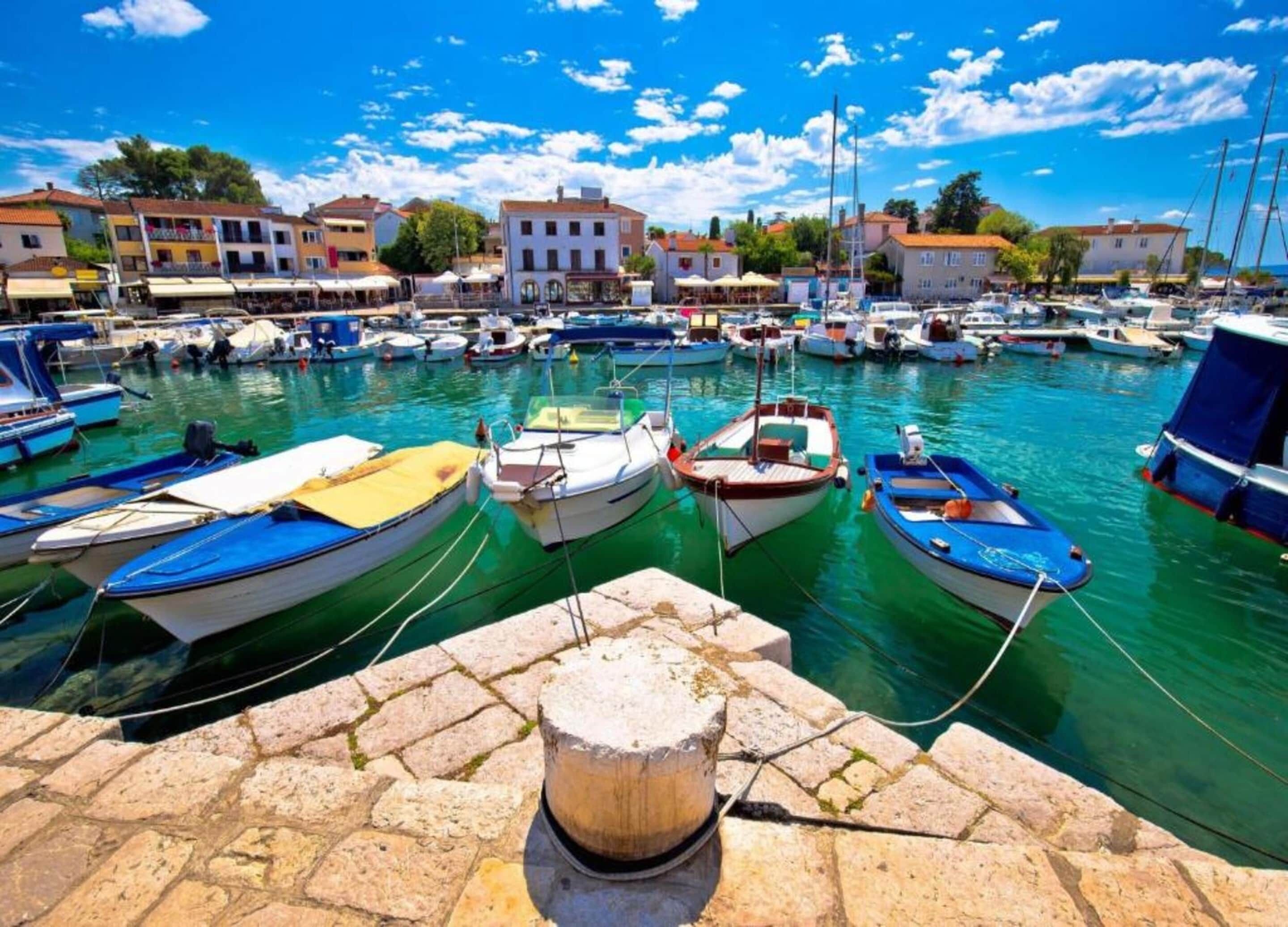 Uma marina com vários barcos atracados, um muro de pedra em primeiro plano e edifícios ao fundo.