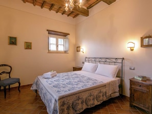 A bedroom with a bed, a chair, a nightstand, a wall-mounted light, and a window.