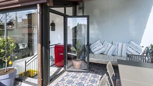 Un petit balcon avec une porte vitrée, une plante en pot, une poubelle rouge et un canapé avec des coussins à rayures.