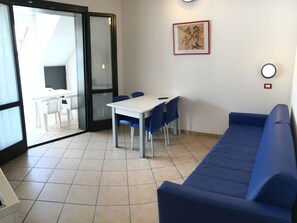 A room with a blue sofa, a white table with blue chairs, a framed picture on the wall, and a sliding glass door leading to a balcony.