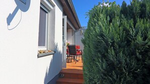 Un estrecho pasillo exterior con pared blanca, ventana y puerta que da a un patio con mesa y sillas.
