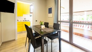 Un espace repas moderne avec une table en verre et des chaises noires, une cuisine avec des meubles jaunes et un balcon doté d’une balustrade.